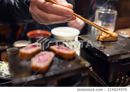 Hand using chopsticks to place topping on gyukatsu beef cooking on a tabletop stone 124824970