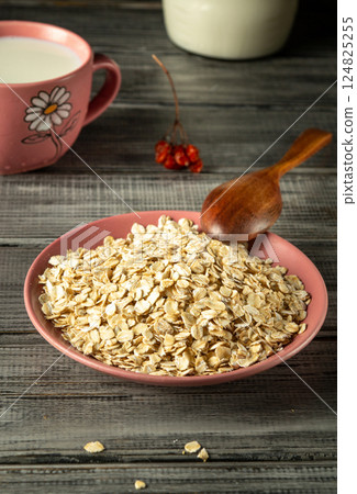 A bowl filled with rolled oats sits on a rustic wooden table, accompanied by a wooden spoon. Nearby, a cup of milk adds to the breakfast vibe, enhancing a calm morning atmosphere 124825255