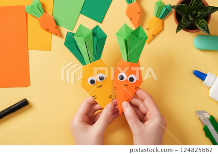 Making a simple origami Easter craft. Hands hold finished crafts of smiling carrots. Around the paper creativity tools. Making a simple origami Easter craft. Hands hold finished crafts of smiling carrots. Around the paper creativity tools. 124825662