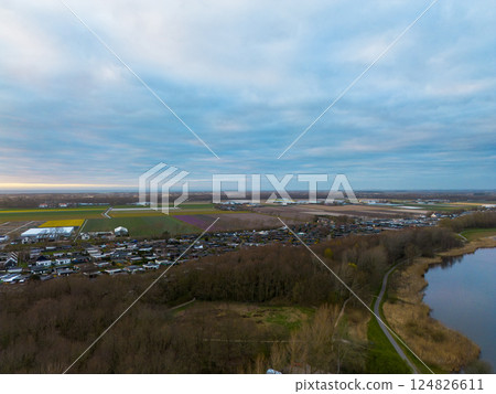 A scenic aerial view of a peaceful rural landscape at dawn, featuring farmlands, colorful flower fields, a quiet village, forested areas, and a calm lake under a cloudy sky. A scenic aerial view of a peaceful rural landscape at dawn, featuring farmlands, colorful flower fields, a quiet village, forested areas, and a calm lake under a cloudy sky. 124826611