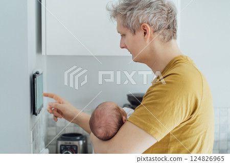 Young man with newborn baby adjusts settings, switch on wall-mounted thermostat or smart control panel, managing temperature, air conditioning system. Modern technology, controlled home environment 124826965