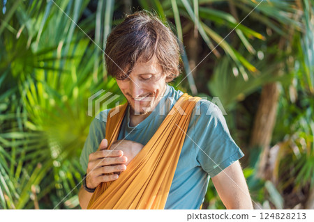 Father holding his little baby in a yellow sling in the park. Warm and loving family moment. Babywearing, fatherhood, and parent-child bonding concept Father holding his little baby in a yellow sling in the park. Warm and loving family moment. Babywearing, fatherhood, and parent-child bonding concept 124828213