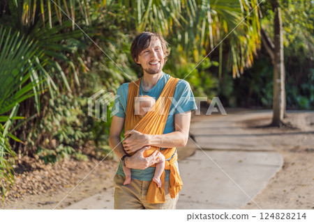 Father holding his little baby in a yellow sling in the park. Warm and loving family moment. Babywearing, fatherhood, and parent-child bonding concept Father holding his little baby in a yellow sling in the park. Warm and loving family moment. Babywearing, fatherhood, and parent-child bonding concept 124828214