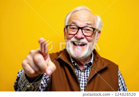 An elderly man wearing glasses and a checkered shirt smiles widely while pointing playfully. His warm expression radiates joy set against a vibrant yellow backdrop. 124828520