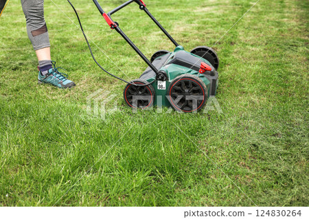 Gardening work tools. Cropped photo of gardener using modern electric aerator-scarifier for cultivation and compaction green grass lawn at backyard. Seasonal landscaping design work. Grass maintenance 124830264