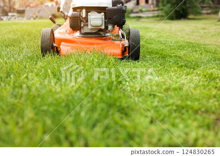 Gardening work tools. Wide angle view of orange electric lawn mower, wheels, motor on bright lush green grass at backyard. Rotary lawn mower machine cut lawn. Professional lawn care service. Gardening work tools. Wide angle view of orange electric lawn mower, wheels, motor on bright lush green grass at backyard. Rotary lawn mower machine cut lawn. Professional lawn care service. 124830265