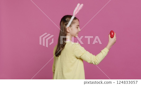 Enthusiastic small child spinning around in front of camera, presenting her red painted egg for easter sunday festivity. Cheery toddler with bunny ears twirling and doing a pirouette. Camera A. 124833057