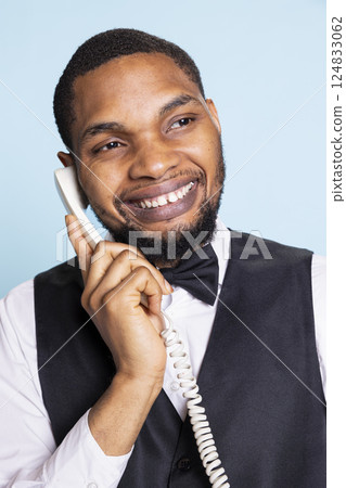 African american hotel porter provides crucial lodging information via landline phone call, discussing about a room reservation with guests. Bellhop answering questions remotely. African american hotel porter provides crucial lodging information via landline phone call, discussing about a room reservation with guests. Bellhop answering questions remotely. 124833062