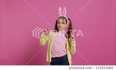 Lovely sweet girl posing in studio with fingers crossed and hoping to receive gifts for easter holiday celebration. Cheerful young child with bunny ears asking for good luck and fortune. Camera B. Lovely sweet girl posing in studio with fingers crossed and hoping to receive gifts for easter holiday celebration. Cheerful young child with bunny ears asking for good luck and fortune. Camera B. 124833068