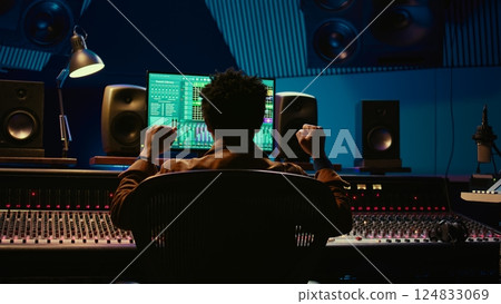 African american sound engineer feeling happy with his recorded song on mixing console, working in music industry post production. Audio technician making a hit song in control room. Camera B. 124833069