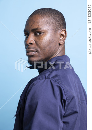 Portrait of african american security officer poses against blue background, looking over his shoulder in studio. Serious guarding personnel dressed in uniform, patrol duty. 124833082