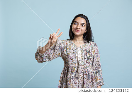 Jolly indian person doing peace sign victory gesture with her hand, studio background on camera. Upbeat woman feeling confident and celebrating win, smiling and being joyful. Jolly indian person doing peace sign victory gesture with her hand, studio background on camera. Upbeat woman feeling confident and celebrating win, smiling and being joyful. 124833122