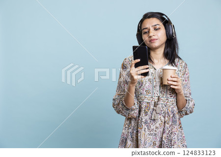 Relaxed happy woman drinking coffee and playing her favorite songs, having fun moving around with her phone. Upbeat indian person dancing on rhythm and serving fresh caffeine drink. Relaxed happy woman drinking coffee and playing her favorite songs, having fun moving around with her phone. Upbeat indian person dancing on rhythm and serving fresh caffeine drink. 124833123