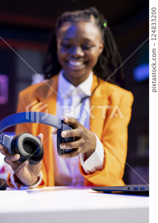 Teleworker putting headphones on to enjoy music while researching data for company project, doing tasks in personal home office. Woman sitting at desk in front of laptop and doing listening session 124833200