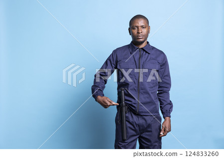 Armed bodyguard showing his baton used for own safety against blue background, pointing at his weapon bat to ensure order and protection. Security agent in uniform on camera. 124833260
