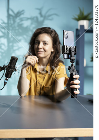 Focus on smartphone attached to selfie stick used by internet star in blurry background to record vlog, chatting with viewers. Close up of phone used by woman to film content for podcasting channel 124833270