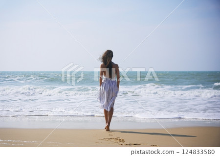 Young woman walking barefoot on beach in summer dress toward calm sea horizon Young woman walking barefoot on beach in summer dress toward calm sea horizon 124833580
