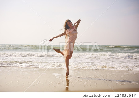 Ballet dancer performs elegant pose on beach. Flowing dress with ocean backdrop Ballet dancer performs elegant pose on beach. Flowing dress with ocean backdrop 124833617