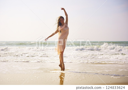 Ballerina dancing on beach in flowing dress with waves and soft morning light 124833624