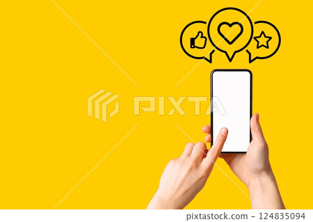 Hand holding a smartphone with interaction icons indicating likes and hearts on yellow 124835094