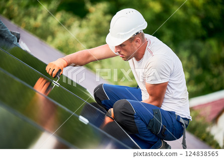 Worker building photovoltaic solar panel system on rooftop of house. Man engineer in helmets and gloves installing solar module with help of hex key outdoors. Alternative energy generation concept. Worker building photovoltaic solar panel system on rooftop of house. Man engineer in helmets and gloves installing solar module with help of hex key outdoors. Alternative energy generation concept. 124838565