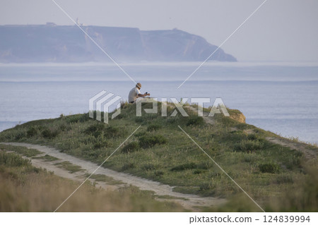 Man relaxing on hilltop overlooking scenic ocean view and using smartphone 124839994