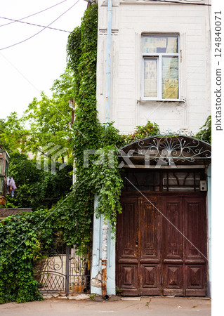 A charming vintage building with a wooden gate and ivy-covered walls, surrounded by lush greenery and rustic details 124840071