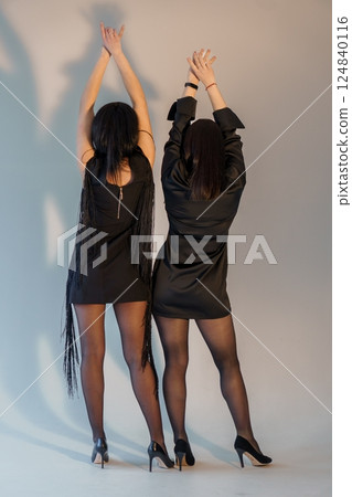 Stylish Women in Black Dresses and High Heels Posing Backward with Raised Arms in Studio 124840116