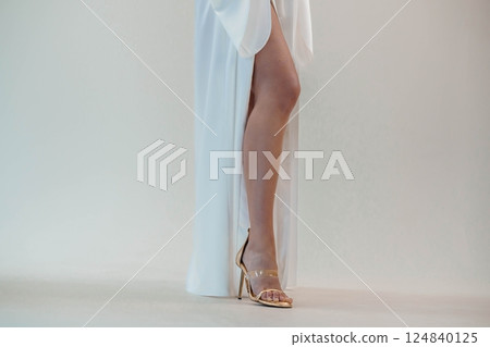 Elegant Female Leg in Gold High Heel Shoe and White Dress on Neutral Background 124840125