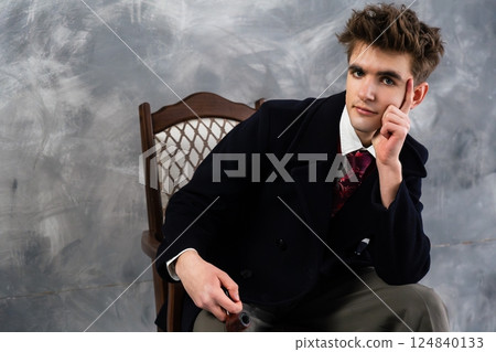 Young elegant man in a dark coat and red tie seated on antique chair, holding pipe and looking thoughtfully at camera 124840133