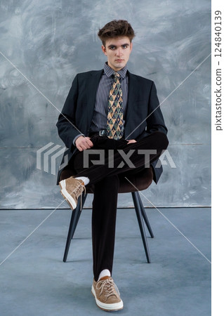 Young man in a dark suit, striped shirt, patterned tie, black pants, and beige sneakers casually seated on chair against gray textured wall Young man in a dark suit, striped shirt, patterned tie, black pants, and beige sneakers casually seated on chair against gray textured wall 124840139