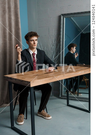 Young man in suit sitting at table with smoking pipe, looking intently forward towards mirror reflection in a modern interior setting Young man in suit sitting at table with smoking pipe, looking intently forward towards mirror reflection in a modern interior setting 124840141