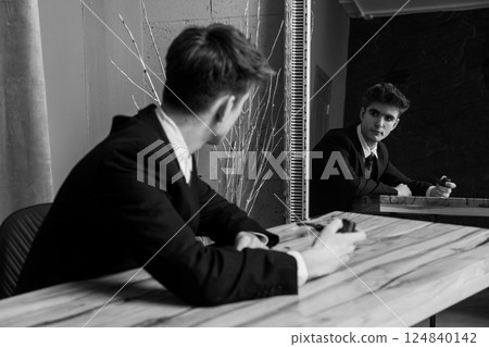Black and white portrait of a man in suit seated at table with pipe, looking into mirror reflecting his image 124840142