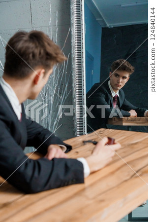 Young man in suit looking at his reflection in mirror while sitting at wooden table, contemplating or assessing himself Young man in suit looking at his reflection in mirror while sitting at wooden table, contemplating or assessing himself 124840144