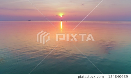 Aerial drone footage capturing a breathtaking tropical sunset reflecting on the calm ocean water surface, creating a mesmerizing colorful gradient from orange and pink to turquoise and green 124840689