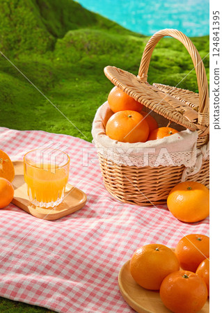 Picnic style bamboo basket with handles containing ripe oranges, placed on red and white checkered carpet, spread on artificial green grass. Oranges placed on wooden plates and scattered around. 124841395