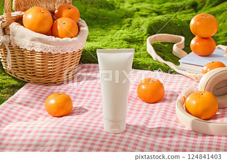 A checkered mat spread on the green lawn of the meadow with many picnic items like mesh bag, headphone, notebook and bamboo basket filled with oranges. Mockup cosmetic tube for designing.  124841403