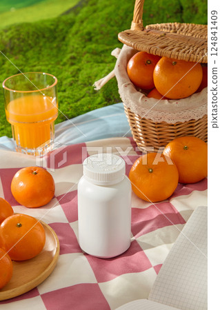 Unlabeled medicine bottles displayed on a picnic rug, spread on lush artificial turf. Oranges peeking out from bamboo baskets. Citrus burst oranges on wooden plates and scattered around the rug.  124841409