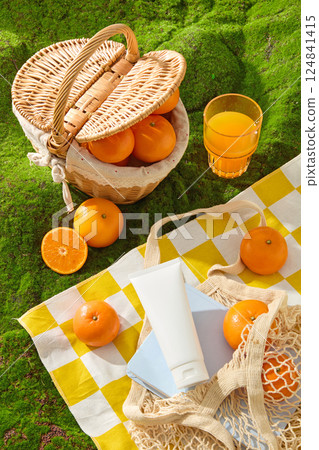 Bright yellow and white checkered picnic rug spread on green grass. Mesh bag containing notebook, unlabeled tube, fresh oranges. Glass of freshly picked orange juice. Shot from above, flat lay. 124841415