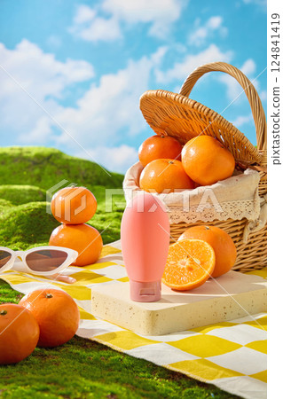 Unlabeled pink cream tube on rectangular pedestal with fruits. Picnic concept with checkered rug spread on green lawn. Fresh oranges filled in a wicker basket. Sunglasses and oranges stacked 124841419