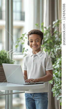 Smiling haired curly african american schoolboy 12 years old is standing and engaged in online learning with laptop and wireless headphones at home. Smiling haired curly african american schoolboy 12 years old is standing and engaged in online learning with laptop and wireless headphones at home. 124842086