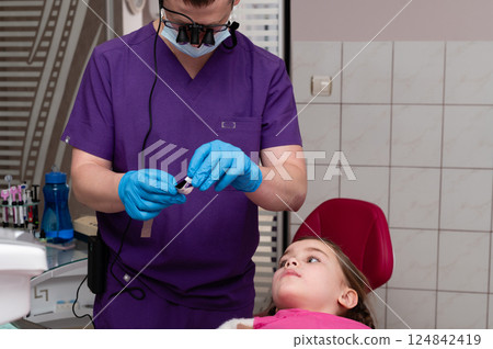 Young girl receiving dental care in a modern clinic with a professional smiling dentist wearing gloves and goggles, ensuring comfort during the procedure 124842419