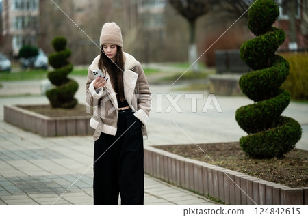 Young woman checks her phone while wearing a stylish winter outfit in a modern urban park adorned with neatly trimmed bushes on a cool day Young woman checks her phone while wearing a stylish winter outfit in a modern urban park adorned with neatly trimmed bushes on a cool day 124842615