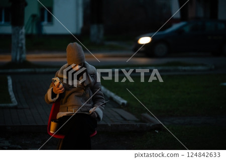 Woman seated on red chair in evening light, engaged with smartphone while reading notepad, surrounded by peaceful urban setting and passing car Woman seated on red chair in evening light, engaged with smartphone while reading notepad, surrounded by peaceful urban setting and passing car 124842633