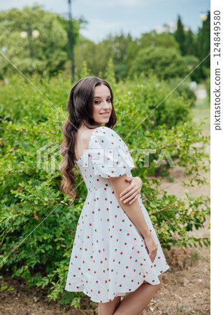 Beautiful slim brunette woman in summer white dress walking among trees in park 124849580