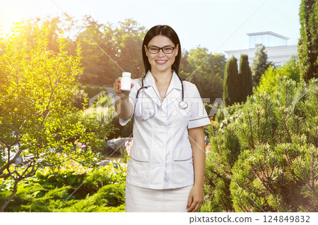 Asian cheerful female doctor is giving an white blank bottle of pills. Physician is advertising meds standing outdoors in the garden. Asian cheerful female doctor is giving an white blank bottle of pills. Physician is advertising meds standing outdoors in the garden. 124849832