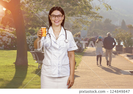 Portrait of a young cheerful female doctor is giving an orange can of pills. Sunny park background. 124849847