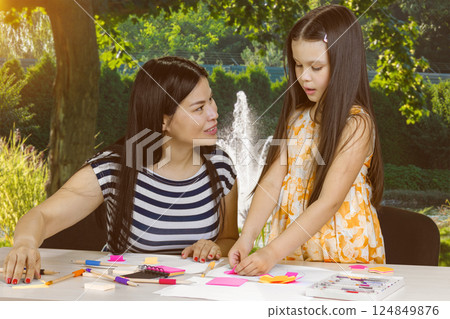Young asian teacher and little girl making paper cutouts. Green summer garden background. Young asian teacher and little girl making paper cutouts. Green summer garden background. 124849876