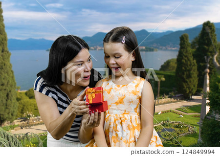 Surprised mother and her littler girl is opening a red gift box. Summer resort landscape background. 124849994
