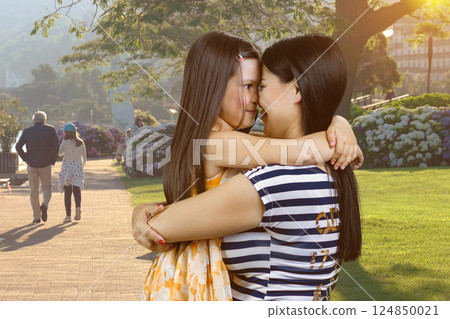 Young brunette mother is hugging her little girl outdoors. City park background. Young brunette mother is hugging her little girl outdoors. City park background. 124850021